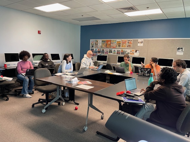 A classroom meeting with people seated around a large rectangular arrangement of tables, using laptops and engaging in discussion. Computer workstations sit along the back wall with posters displayed above them.