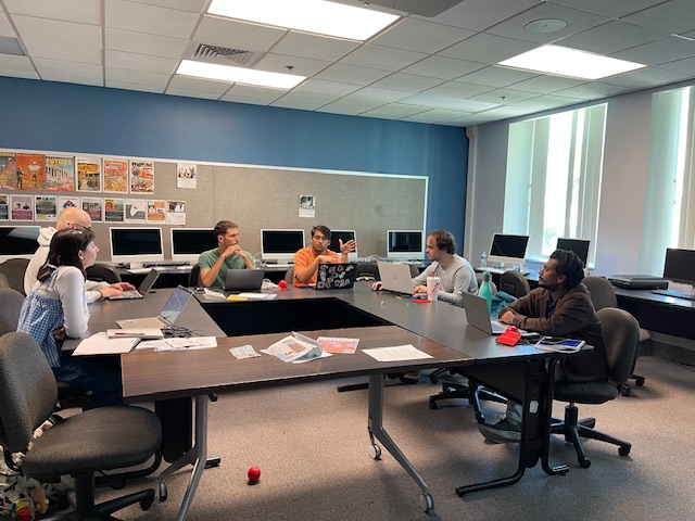 A classroom scene with several people seated around a large U‑shaped table, working on laptops and discussing. Computer monitors line the back wall, and printed posters are pinned to a bulletin board.