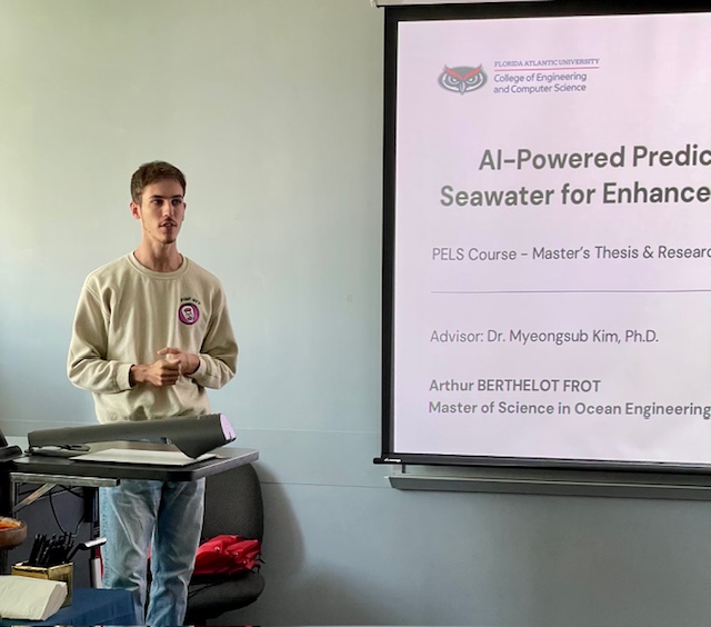 A person standing at the front of a classroom next to a podium, presenting a slide titled “AI‑Powered Prediction of Seawater for Enhanced…” with academic information and university branding visible.