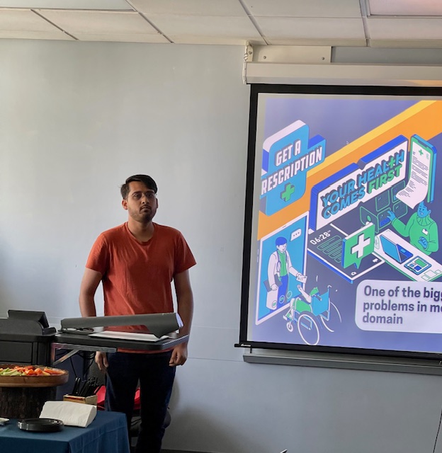 A person standing near a podium presenting to a group, with a projected slide showing colorful graphics about healthcare, prescriptions, and medical services.