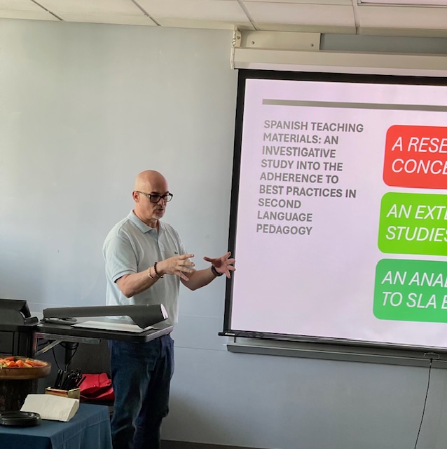 A presenter gesturing beside a podium while a classroom projection screen displays a slide about Spanish teaching materials, educational research, and second‑language pedagogy.