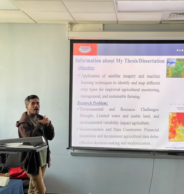 A presenter beside a podium giving a talk while a slide projected on the screen details thesis or dissertation information, including objectives on satellite imagery and agricultural research challenges.