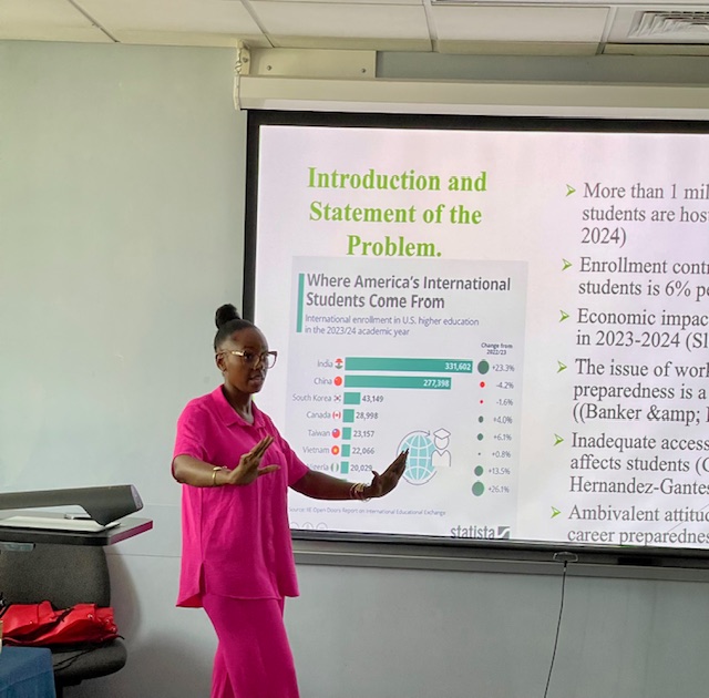 A person giving a presentation while a projection screen shows a slide titled “Introduction and Statement of the Problem,” alongside a chart about where international students come from and related educational data.