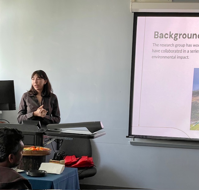 A person giving a presentation in a classroom, standing beside a podium while a projection screen displays a slide titled “Background” with text about research collaboration and environmental impact.