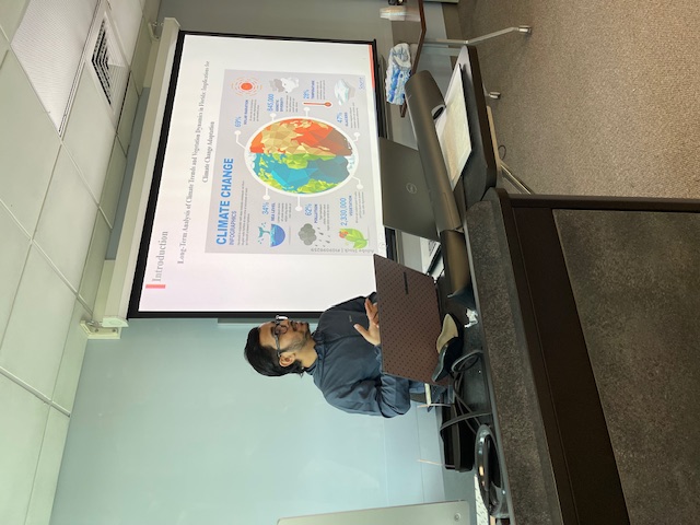 A person sits at a computer desk in a classroom, speaking during a presentation.