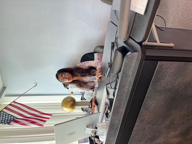 A presentor sits at a computer desk in a classroom, speaking during a presentation.