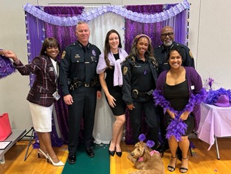 A photo of six people including officers and staff wearing black and purple clothing standing in front of a purple backdrop for Domestic Violence Awareness. On the floor in front of the people is a tan colored dog wearing a purple headband.