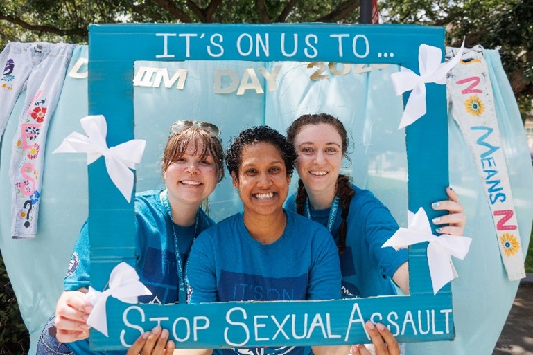 Three individuals are holding up a teal pledge sign that is in a frame that they placed in front of them that says, “it’s on us to stop sexual assault.” Behind them is a teal backdrop with two pairs of jeans hanging on either side. In the middle of the jeans, there is a sign that says Denim Day. 