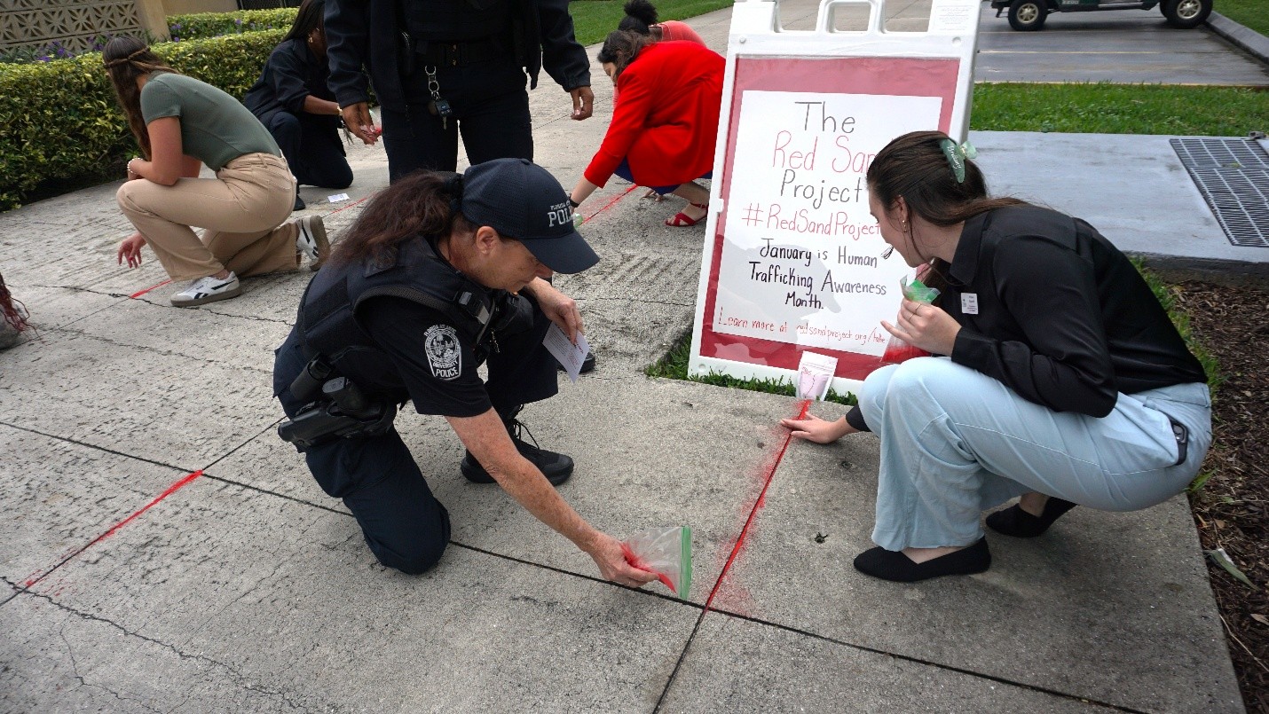 Staff, students and officers are placing red sand on the sidewalk. They are kneeling and placing the sand in between the cracks of the sidewalk. There is a sign that says “Red Sand Project” to raise awareness about Human Trafficking. 