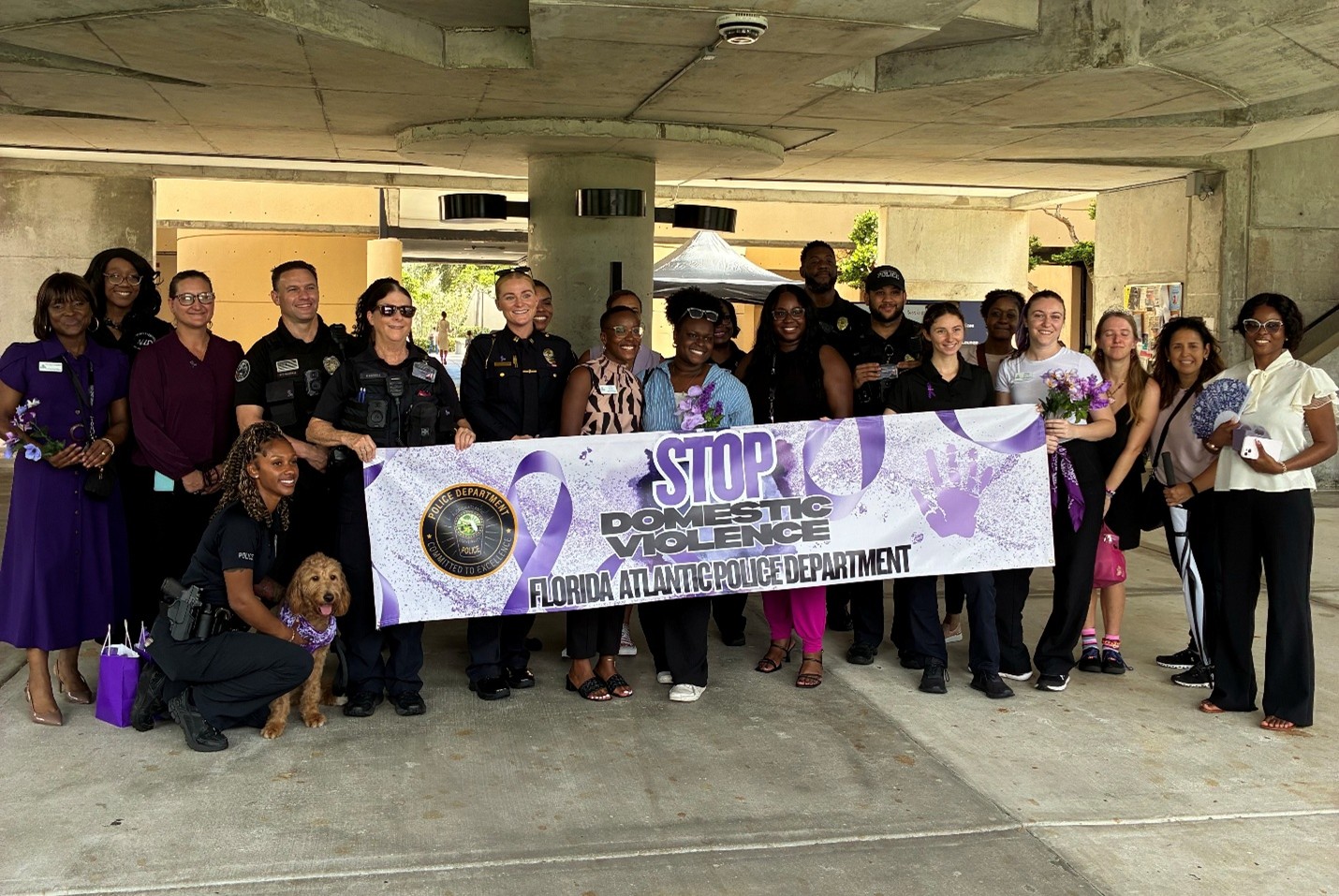 A photo of staff, students and officers standing behind a banner. The banner says “ stop domestic violence Florida Atlantic Police Department.” People are wearing purple, there is purple is in the banner because purple is the official color for Domestic Violence. 