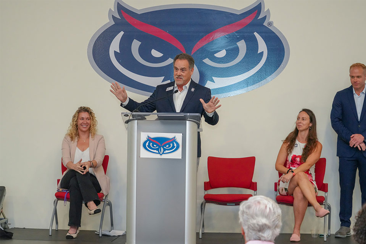 A man in a suit and tie delivers a speech at a podium during a formal event with the FAU owl logo affixed to a white wall in the background.