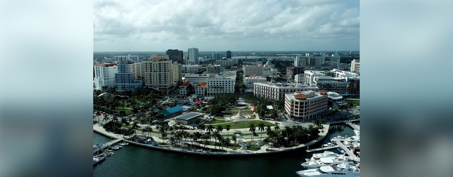 Aerial view of the West Palm Beach waterfront featuring a curved green park lined with palm trees, city buildings, and luxury yachts docked in a marina.