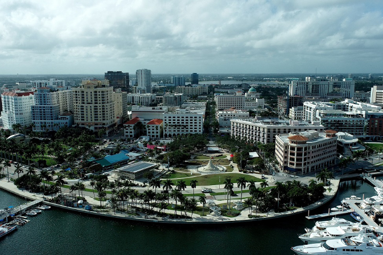 Aerial view of the West Palm Beach waterfront featuring a curved green park lined with palm trees, city buildings, and luxury yachts docked in a marina.
