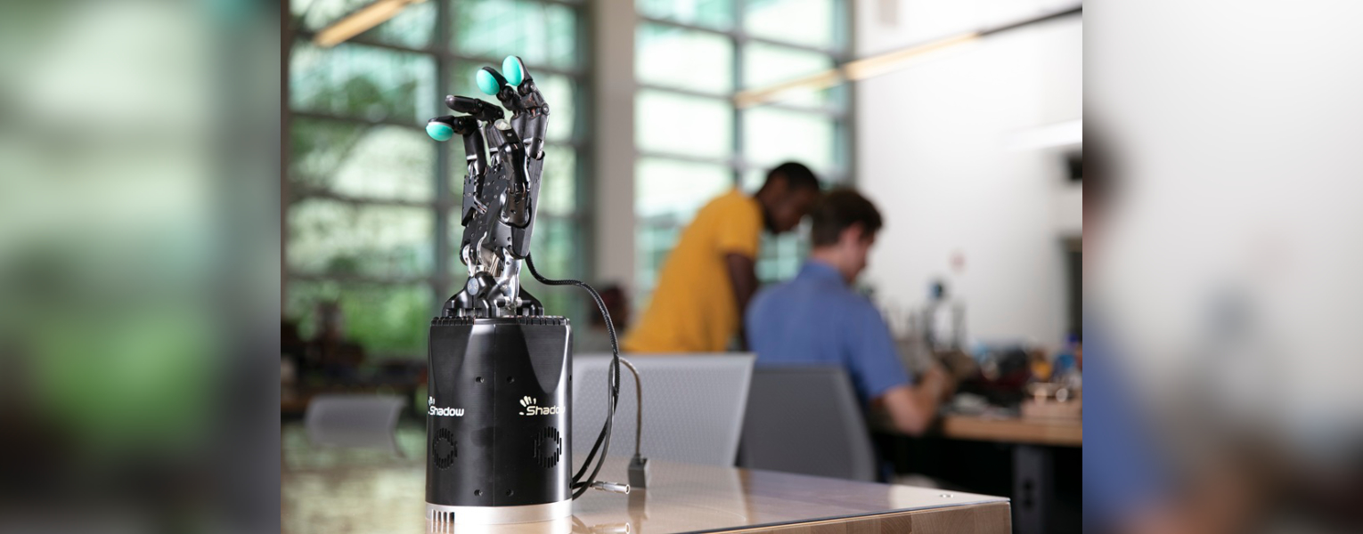 A black Shadow robotic hand with teal fingertips standing on a reflective table inside a blurred laboratory with two people working in the background. (Photo by Alex Dolce)