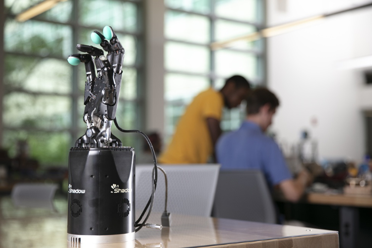 A black Shadow robotic hand with teal fingertips standing on a reflective table inside a blurred laboratory with two people working in the background.
