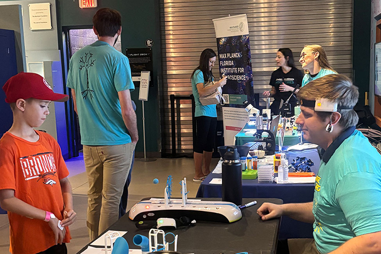A child wearing a red cap and orange T-shirt stands at a table facing a seated man in a teal “Neuro Squad” shirt, who has a white sensor headband on his forehead. Between them is a small device with buttons and upright components. In the background, several people gather around a booth with a “Max Planck Florida Institute for Neuroscience” banner, along with microscopes and science materials.