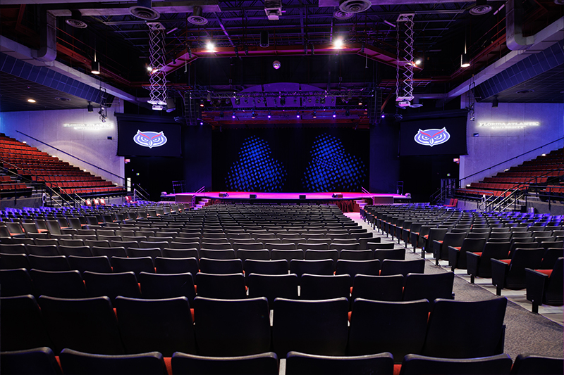 Front-facing view of the Carole and Barry Kaye Auditorium with rows of theater seating leading to a large stage, overhead lighting rigs, and two screens displaying the Florida Atlantic owl logo on either side of the stage