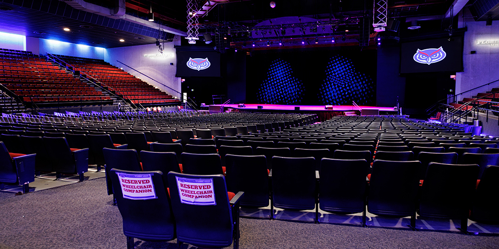 Interior view of the auditorium with tiered seating and a large stage illuminated by purple and blue lighting. Reserved wheelchair companion seating is visible in the foreground, and screens display the FAU owl logo.
