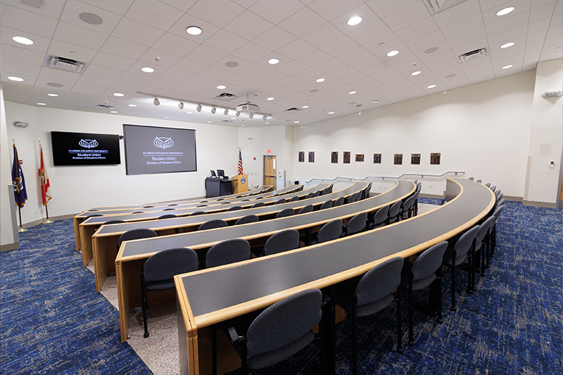 House Chambers featuring tiered, curved desk seating arranged in a lecture-style format, blue carpeted flooring, podium at the front, and large display screens showing Florida Atlantic University Student Union branding.