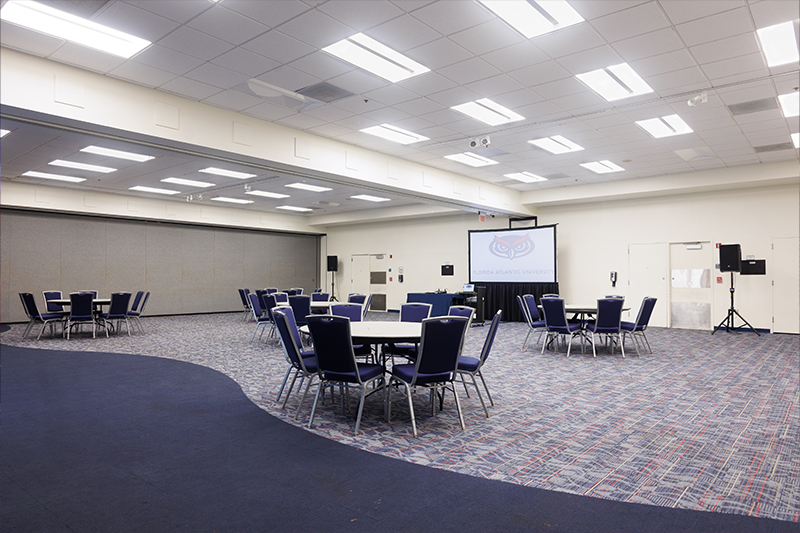 : Live Oak room set for an event, featuring round tables with chairs arranged throughout a large, open room, patterned carpet, neutral walls, and a projection screen at the front displaying the Florida Atlantic logo 