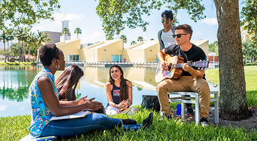 Students sitting on campus lawn reading books and playing guitar